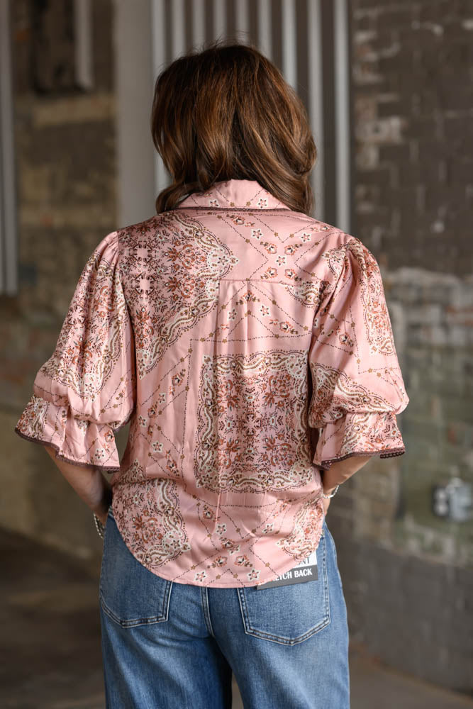 A woman viewed from behind wearing a light pink patterned blouse with ruffled three-quarter sleeves and blue jeans, standing against a rustic brick wall.
