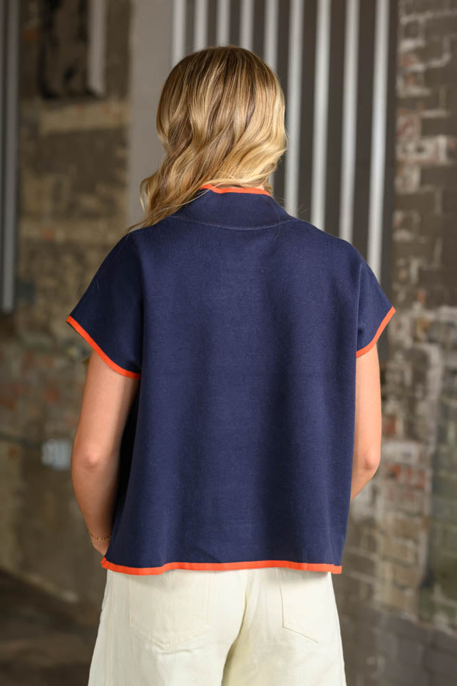 A woman viewed from behind wearing a navy blue short-sleeve knit top with bright orange trim along the collar, sleeve cuffs, and hem, paired with cream pants.