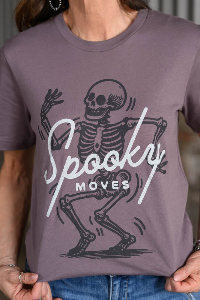 A person wearing a mauve-purple short-sleeve t-shirt with a black and white graphic of a dancing skeleton and the text 'Spooky Moves'.