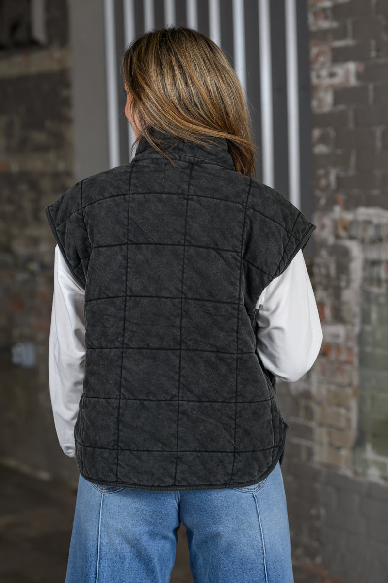 A woman viewed from behind wearing a dark charcoal gray quilted vest over a white long-sleeve shirt and blue jeans, standing in front of a rustic brick wall.