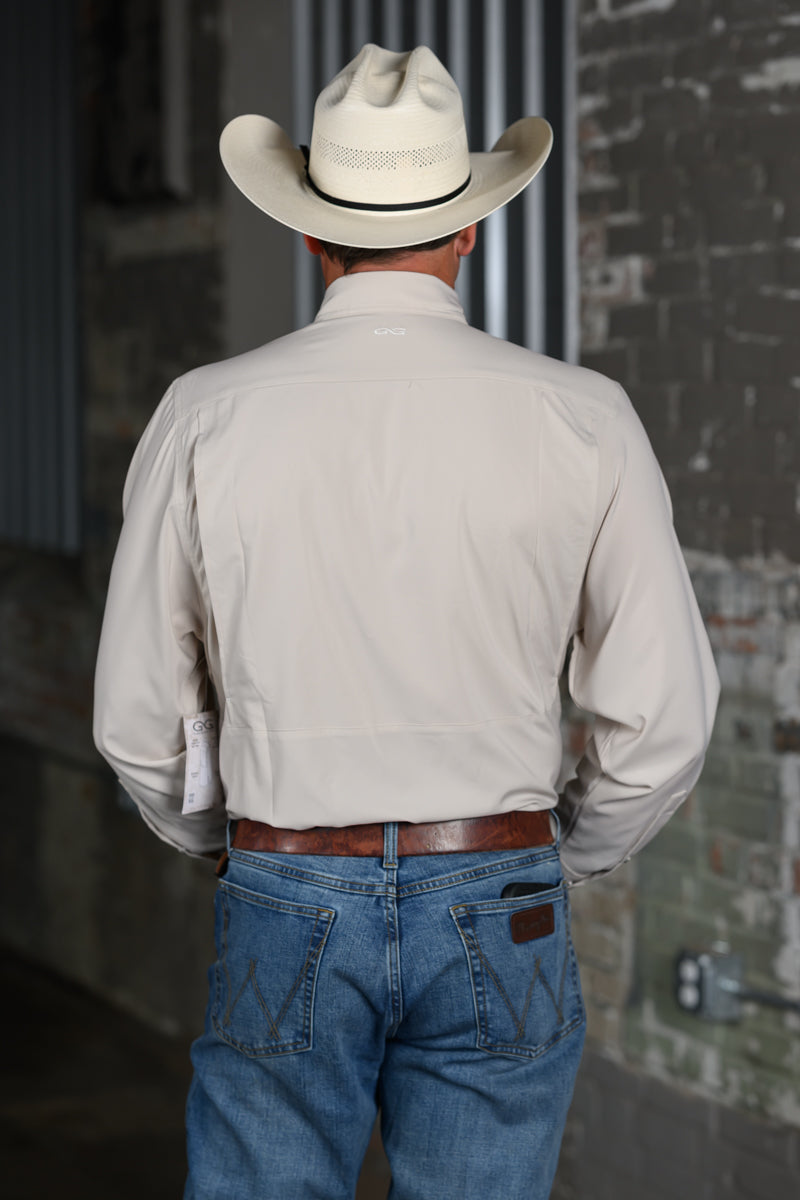 A man viewed from behind wearing a light beige long-sleeve western shirt, a cream cowboy hat, and blue jeans, standing against a rustic brick and metal wall.