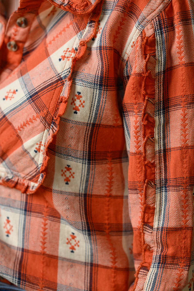 Close-up of a distressed orange plaid shirt with frayed edges, featuring a cream-colored geometric pattern block and brown buttons.