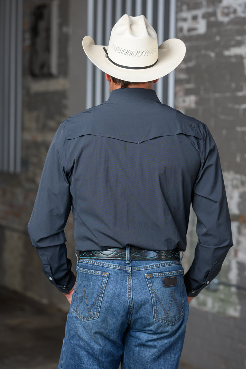 A man viewed from behind wearing a dark slate blue western long-sleeve shirt, blue jeans, and a cream cowboy hat, standing against a rustic brick wall.