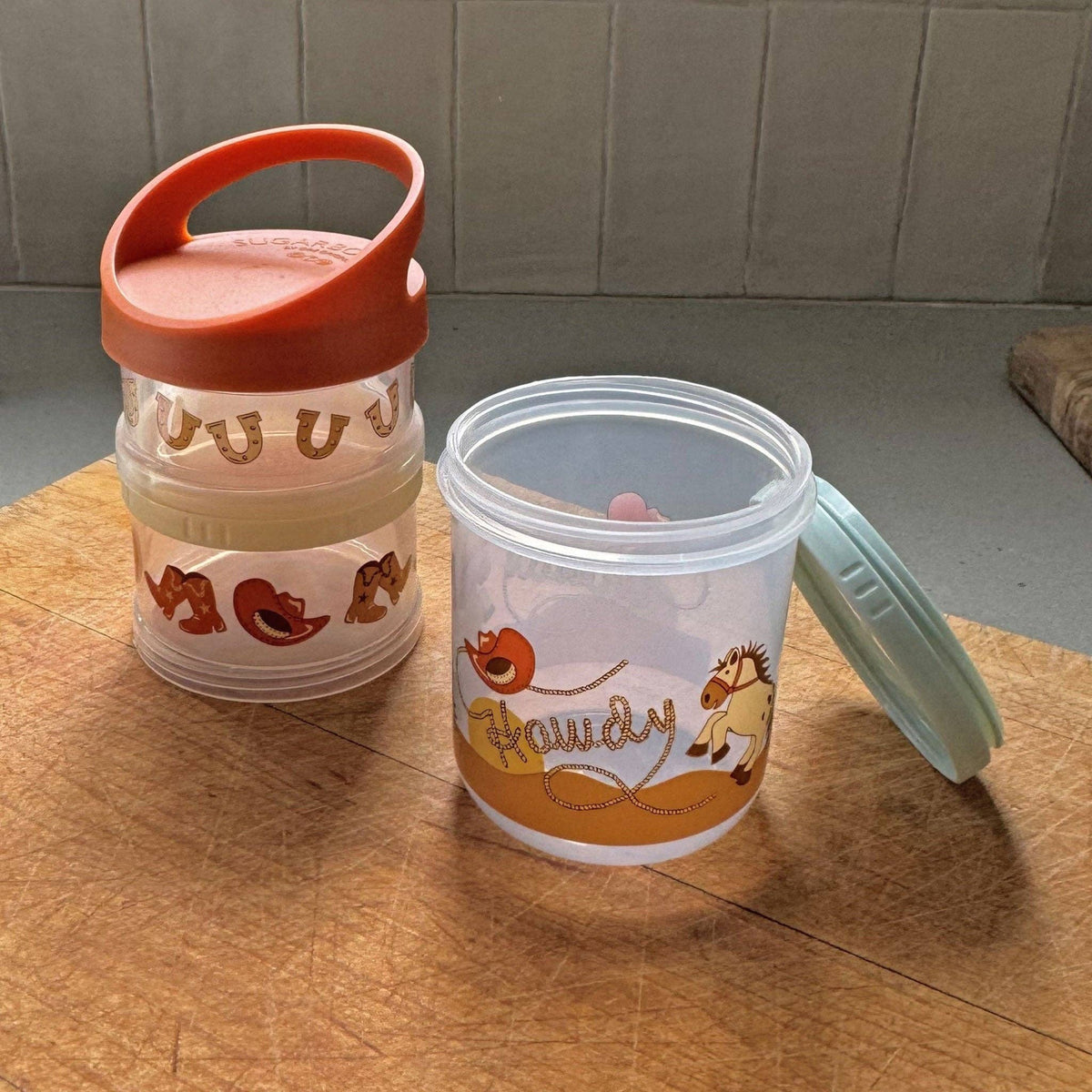 A set of three stacked snack containers with a boots and Howdy motif, in orange, green, and clear colors, with animal illustrations.
