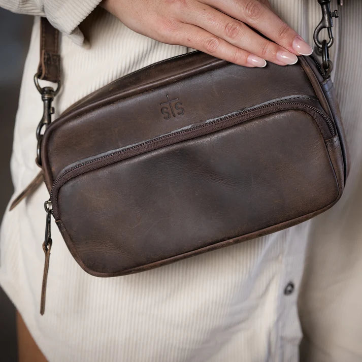 A close-up of a person wearing a dark brown leather crossbody bag with a zipper pocket and embossed logo, shown against a cream ribbed knit top.