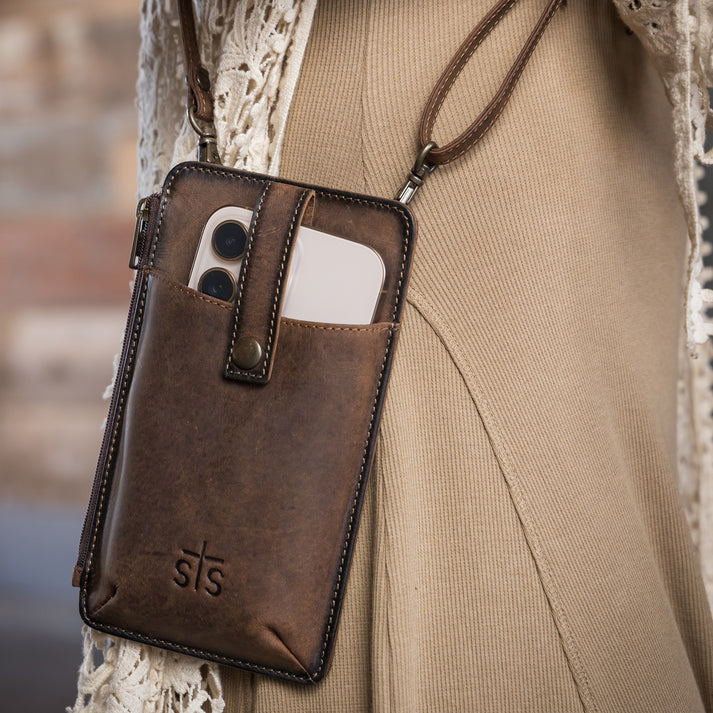 A brown leather phone pouch with a wrist strap, shown worn over a beige ribbed sweater. The pouch has a front pocket holding a smartphone and features an embossed 'sts' logo near the bottom.