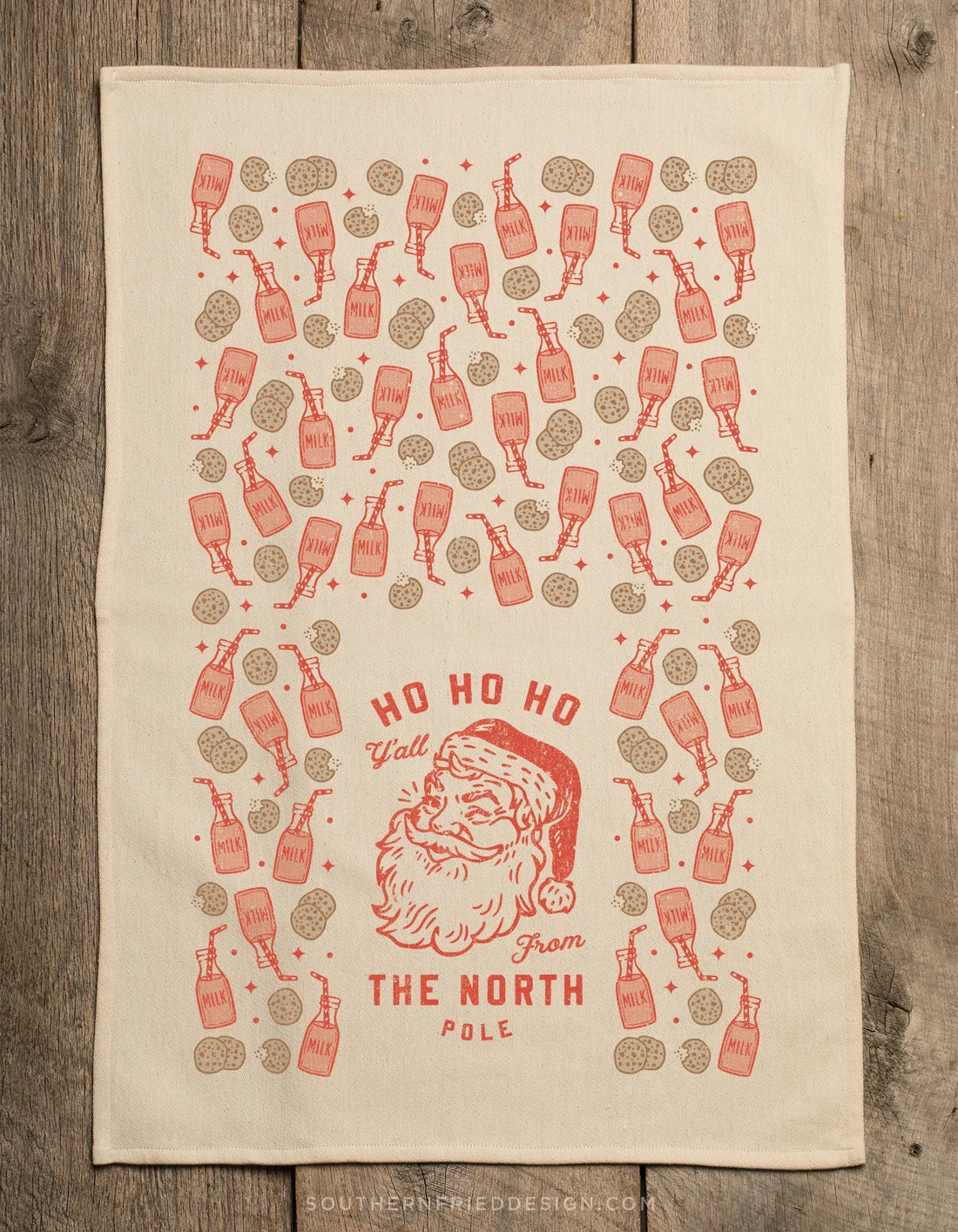 A rectangular cream-colored tea towel with a red and brown holiday print, laid flat on a rustic wooden surface. The design features a central illustration of Santa Claus with the text 'HO HO HO From THE NORTH POLE' surrounded by repeated illustrations of bottles and round brown shapes.