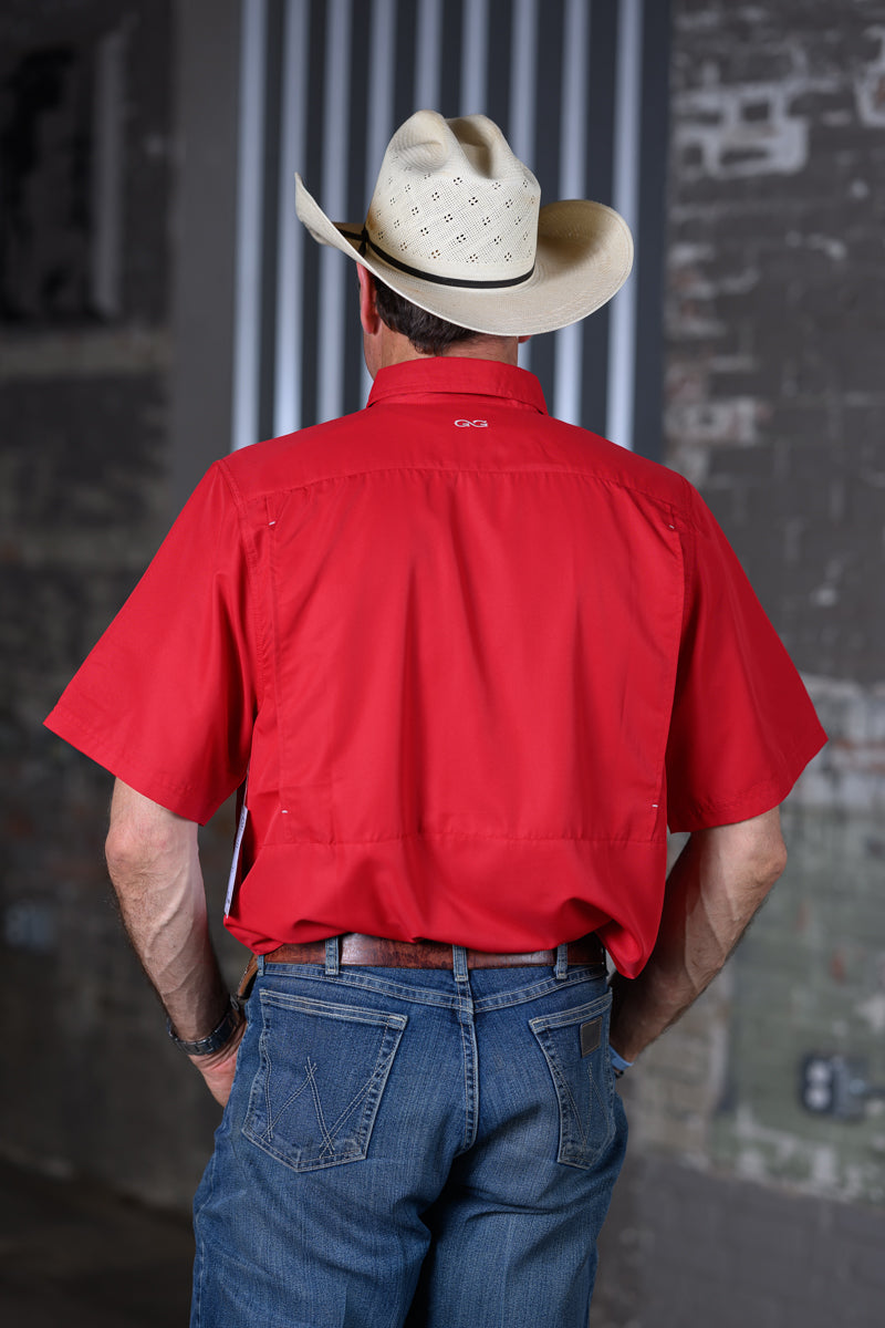 A man viewed from behind wearing a bright red short-sleeve button-up shirt, blue jeans, and a light beige cowboy hat, standing against a gray brick and metal background.