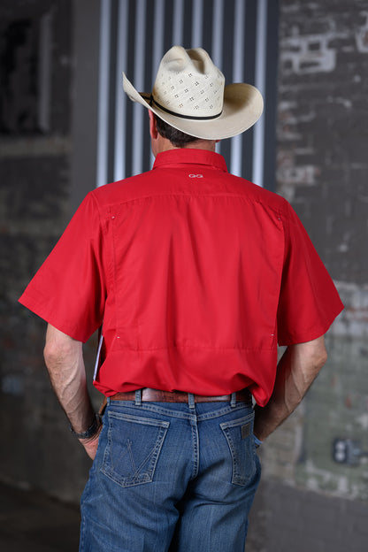 A man viewed from behind wearing a bright red short-sleeve button-up shirt, blue jeans, and a light beige cowboy hat, standing against a gray brick and metal background.