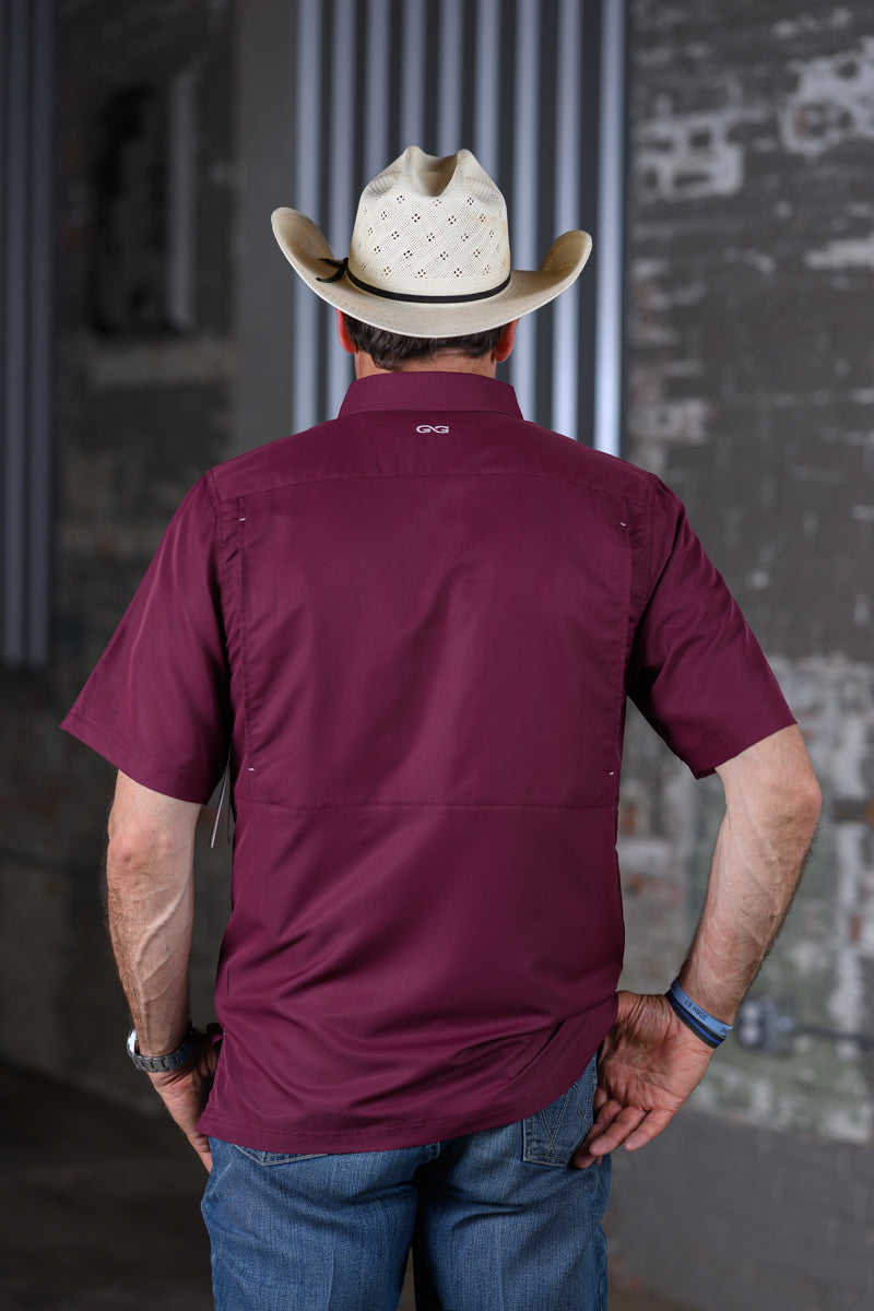 A man viewed from behind wearing a burgundy short-sleeve button-up shirt and a light beige cowboy hat, standing in front of a gray brick wall.