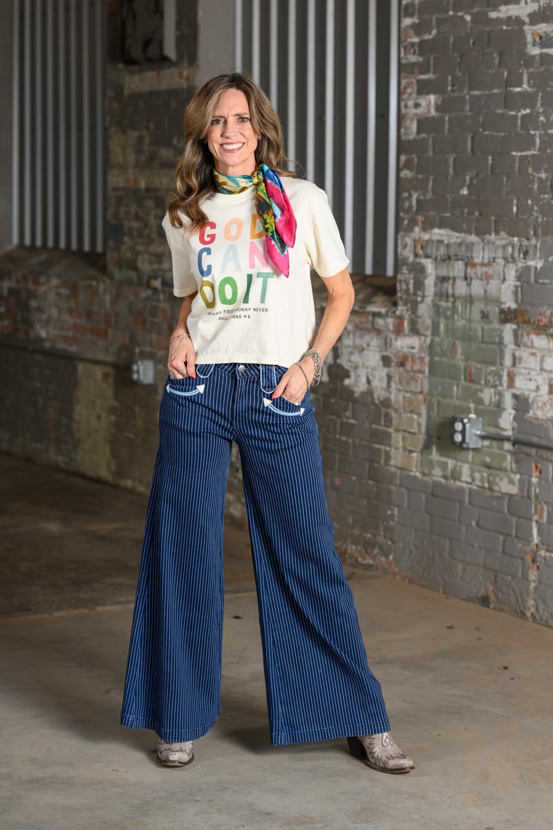 A woman stands in an industrial loft setting wearing a cream graphic t-shirt with colorful text, wide-leg pinstriped blue jeans, and a vibrant multicolor neck scarf.