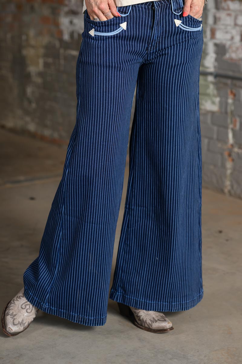 A person wearing dark blue pinstriped wide-leg pants with white contrast stitching on the pocket edges, paired with patterned cowboy boots, standing against a rustic brick wall.