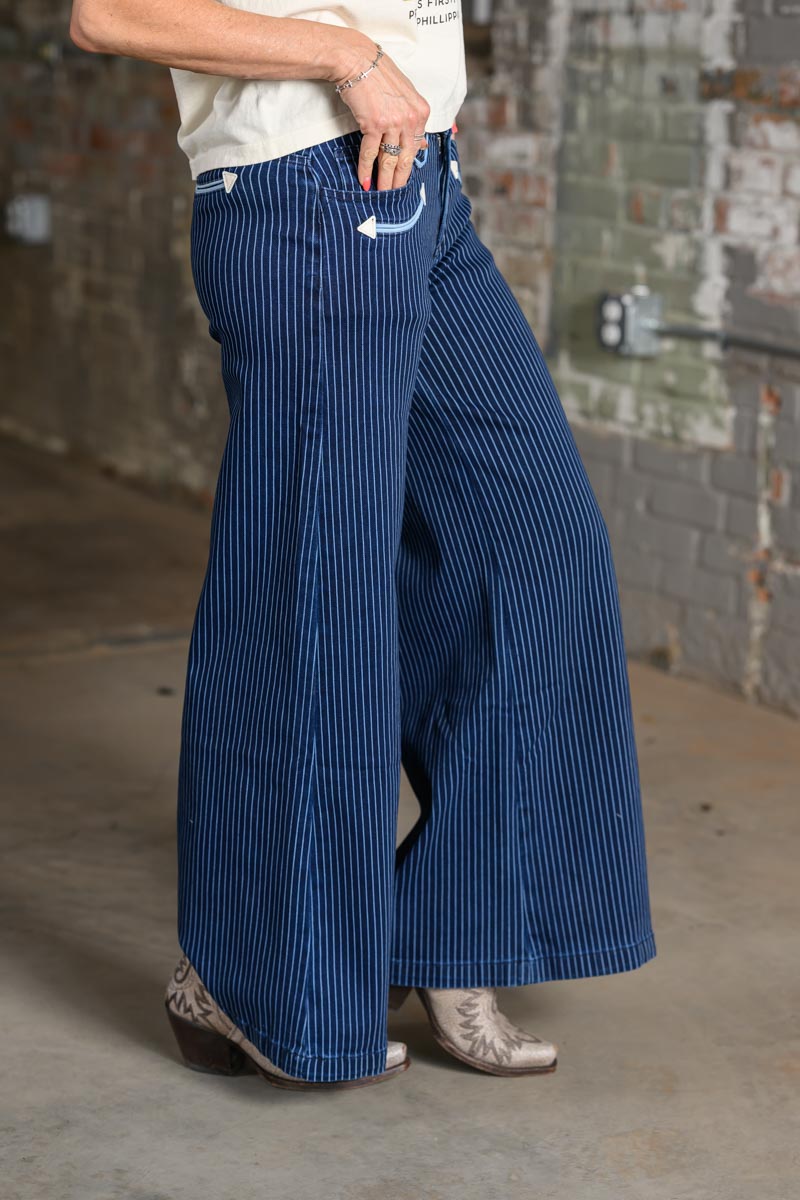 A person wearing wide-leg blue jeans with thin white vertical pinstripes, paired with a white top and western-style boots, standing in front of a rustic brick wall.