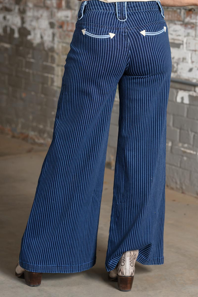 A person shown from the back wearing blue pinstriped wide-leg pants with white decorative stitching on the back pockets, standing in front of a weathered brick wall.