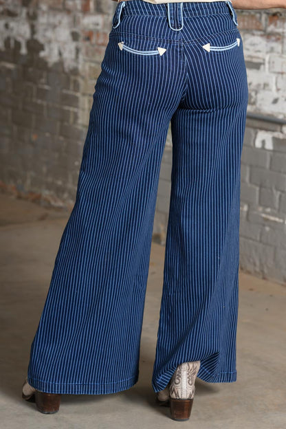 A person shown from the back wearing blue pinstriped wide-leg pants with white decorative stitching on the back pockets, standing in front of a weathered brick wall.