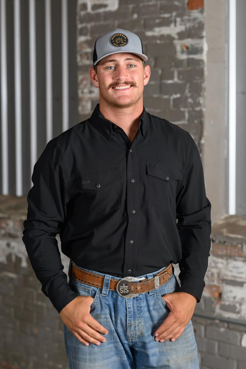A man wearing a black long-sleeve button-down shirt, blue jeans, a brown leather belt, and a gray baseball cap, standing in front of a rustic brick wall.