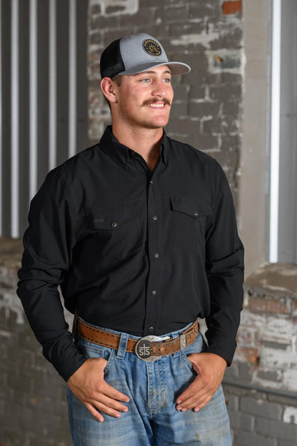 A man wearing a black long-sleeve button-down shirt, blue jeans, a brown leather belt, and a gray and black trucker hat, standing in front of a rustic brick wall.