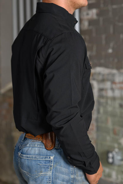 A man wearing a black long-sleeve button-down shirt and blue jeans, shown from a side-back angle against a blurred brick wall background.