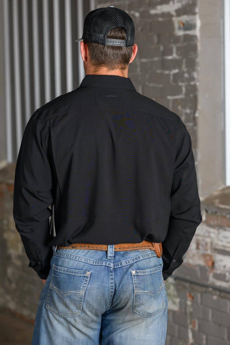 A man viewed from behind wearing a black long-sleeve button-down shirt, blue jeans, a brown leather belt, and a black trucker hat, standing in front of a brick wall.