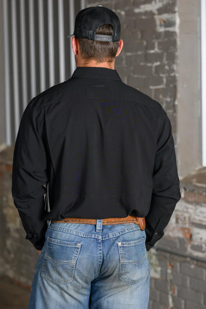 A man viewed from behind wearing a black long-sleeve button-down shirt, blue jeans, a brown leather belt, and a black trucker hat, standing in front of a brick wall.