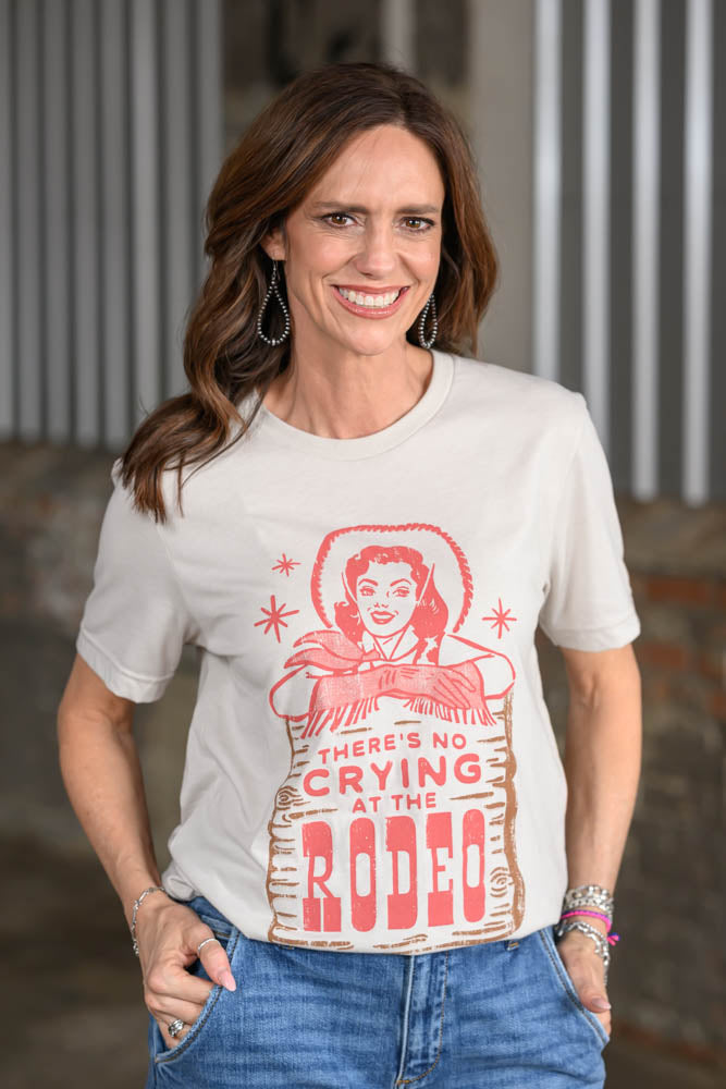 A woman wearing a cream-colored graphic t-shirt with a red and brown western-style print that reads 'THERE'S NO CRYING AT THE RODEO', paired with blue jeans.