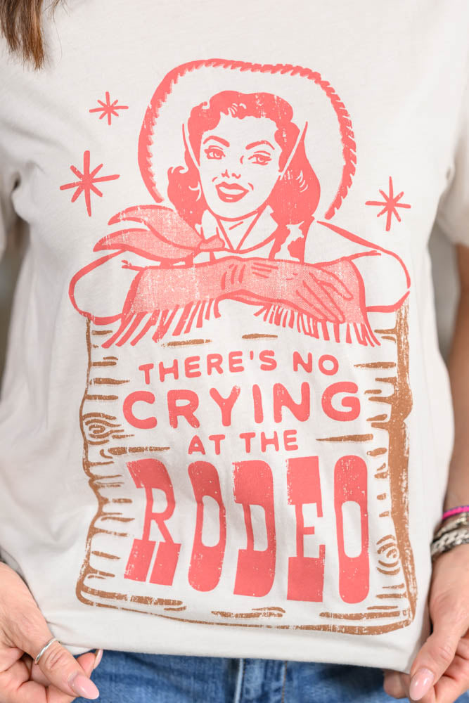 A close-up of a person wearing a white graphic T-shirt with a red and brown vintage-style western print featuring a woman and the text 'THERE'S NO CRYING AT THE RODEO'.