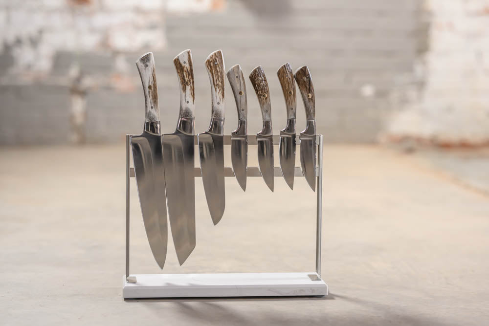 A set of kitchen knives with polished silver blades and textured brown handles, displayed upright in a clear acrylic stand with a white base on a concrete floor.