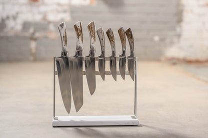 A set of kitchen knives with polished silver blades and textured brown handles, displayed upright in a clear acrylic stand with a white base on a concrete floor.