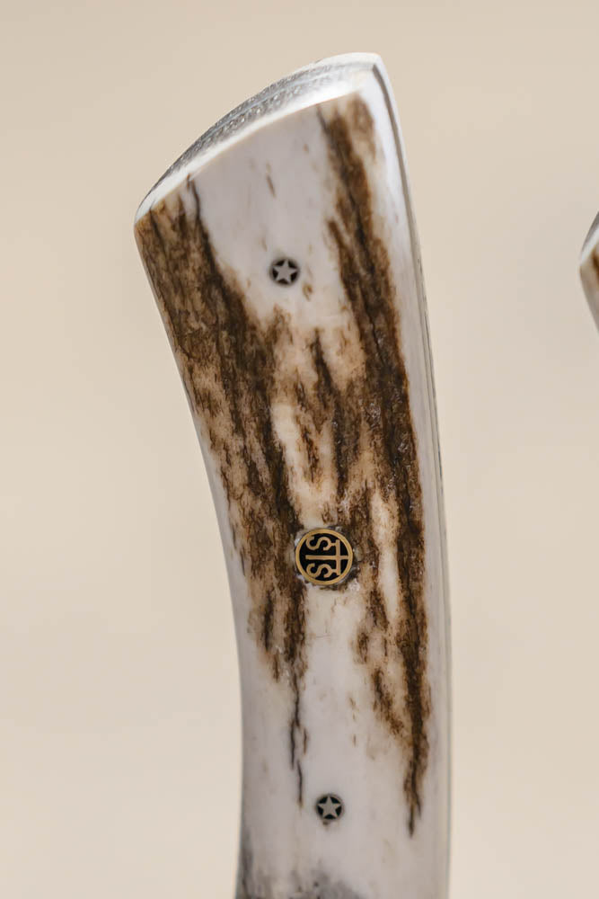 Close-up of a knife handle made of natural antler with dark brown veining, featuring brass and black decorative pins, set against a solid beige background.