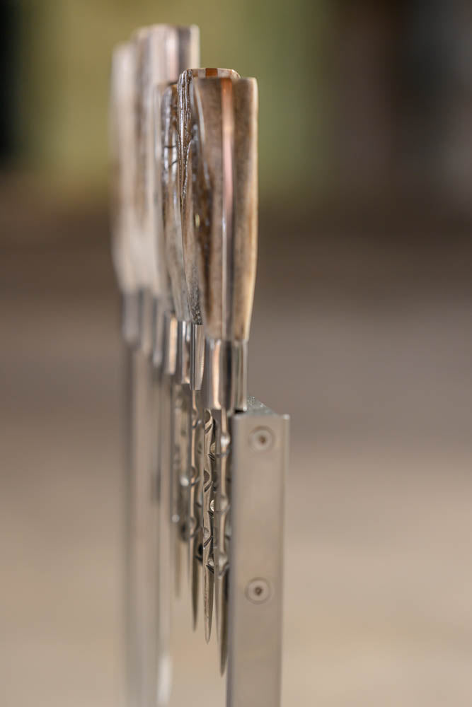 A close-up shot of several knives with ram horn handles standing upright in a metal rack, set against a softly blurred warm-toned background.
