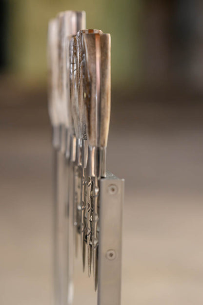 A close-up shot of several knives with ram horn handles standing upright in a metal rack, set against a softly blurred warm-toned background.