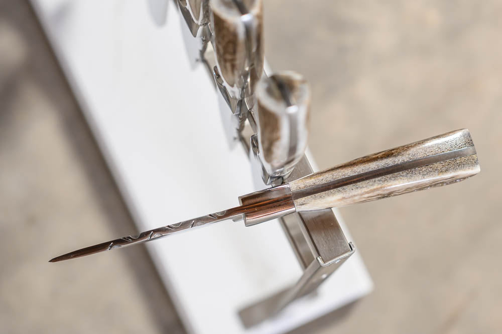 A close-up of a polished metal knife with a textured silver handle resting on a white stand against a blurred gray background.