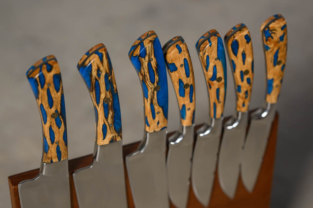 A set of kitchen knives with polished steel blades and custom carved wooden handles featuring a blue and tan spotted pattern, displayed in a wooden block.