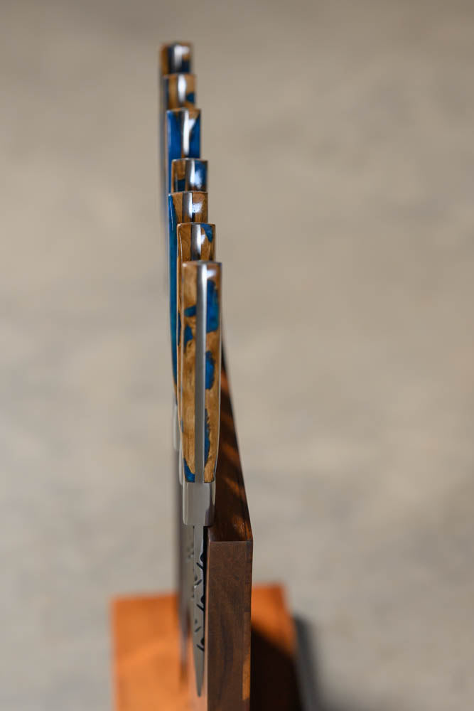 A close-up side view of a set of kitchen knives with blue and silver patterned handles, mounted on a dark wooden block stand against a soft gray background.