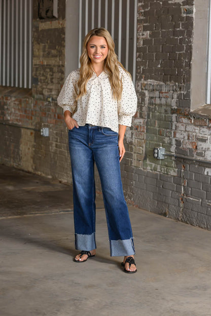 A woman stands in an industrial loft setting wearing a white long-sleeve blouse with small black polka dots, blue wide-leg jeans with rolled cuffs, and black sandals.
