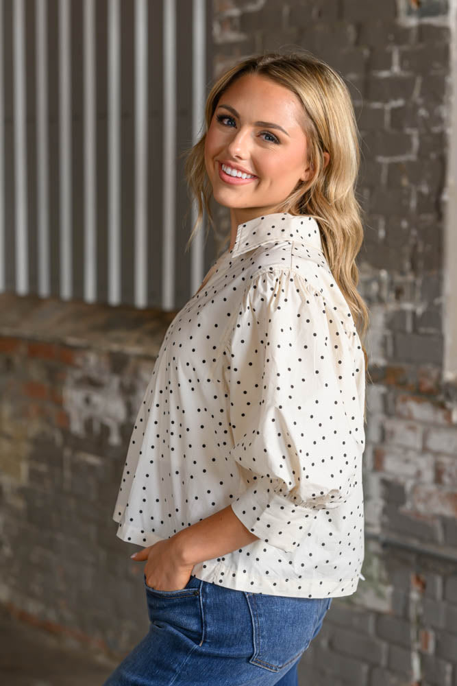 A woman wearing a cream-colored blouse with black polka dots and blue jeans, posing in front of a rustic brick wall and corrugated metal panel.
