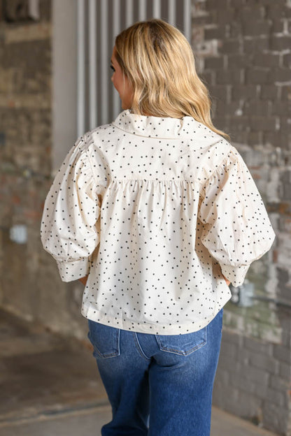 A woman viewed from behind wearing a cream-colored blouse with small black polka dots and voluminous puff sleeves, paired with blue jeans, standing against a rustic brick wall.