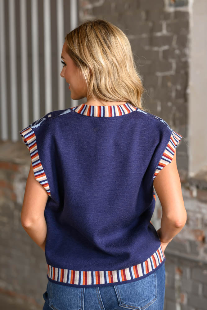 A woman viewed from behind wearing a navy blue knit sweater vest with red, white, and blue striped trim at the collar, sleeve cuffs, and hem, paired with blue jeans.