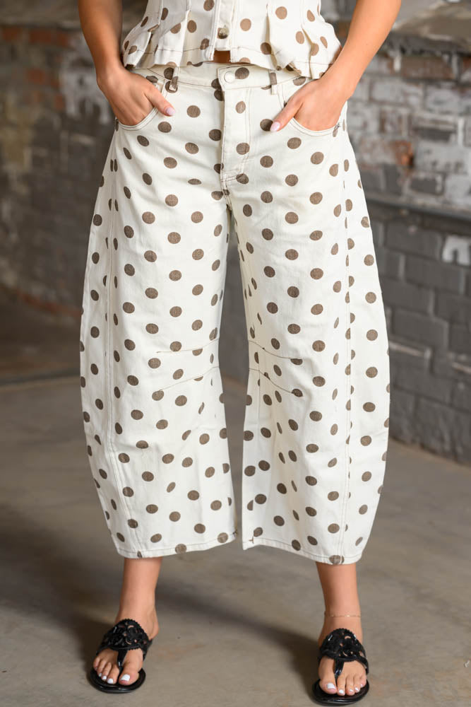 A person wearing wide-leg cropped pants with an off-white base and medium brown polka dots, paired with black thong sandals, standing against a gray brick wall.