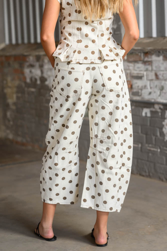 A woman shown from the back wearing a matching sleeveless top and wide-leg cropped pants set in an off-white fabric with brown polka dots, standing against a brick and corrugated metal wall.