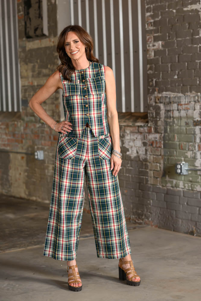 A woman models a sleeveless plaid jumpsuit in a warehouse setting with brick walls. The outfit features a green, red, and white plaid pattern with a button-up front and wide legs.