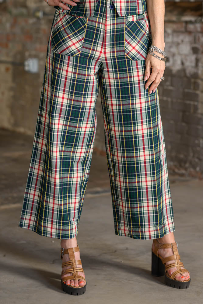 A person wearing high-waisted, wide-leg plaid pants in green, red, and cream, paired with brown strappy platform sandals, standing against a rustic brick wall.