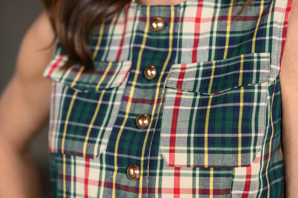 Close-up of a sleeveless plaid top worn by a person, featuring a green, red, yellow, and white tartan pattern with metallic bronze buttons and chest pockets.