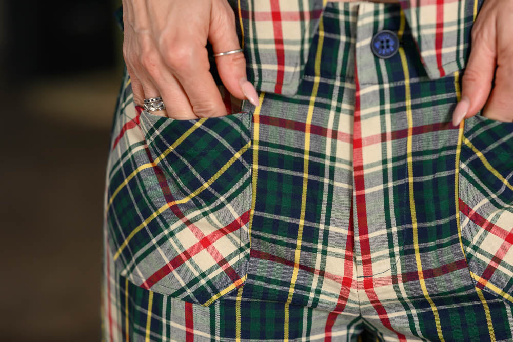 Close-up of a person wearing plaid trousers, with both hands reaching into the front pockets. The fabric has a tartan pattern in green, red, yellow, and cream, with a blue button visible at the waist.