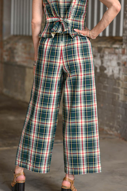 A person wearing plaid wide-leg pants and a matching tie-front top, photographed from the back in an industrial interior with brick and corrugated metal walls.