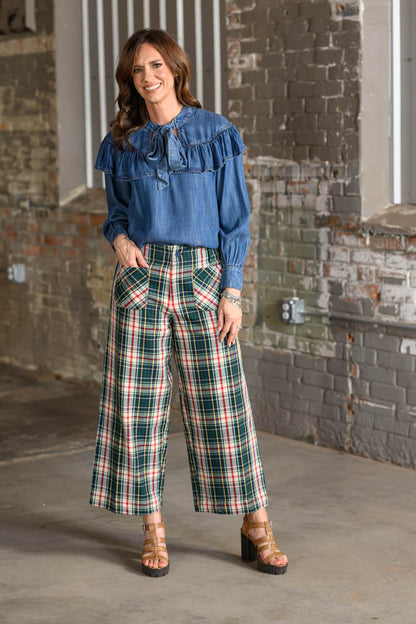 A woman stands in an industrial loft setting with brick walls, wearing a blue ruffled long-sleeve blouse and green, red, and white plaid wide-leg cropped pants with brown block-heel sandals.