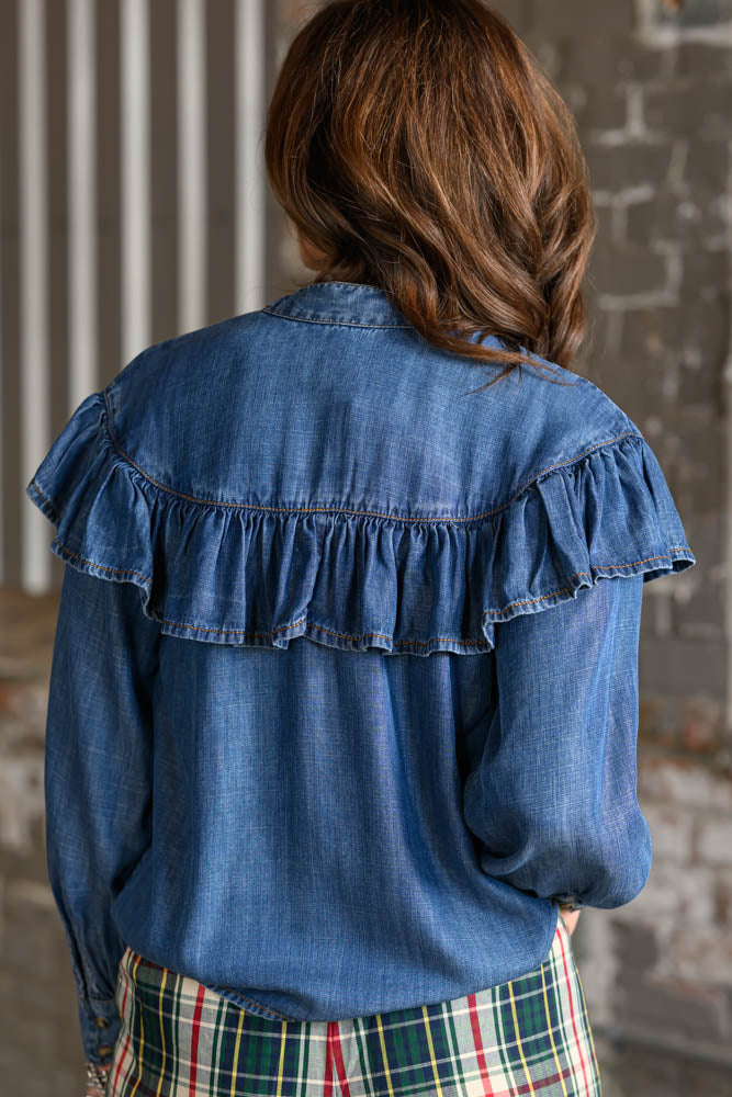 A woman viewed from behind wearing a blue denim long-sleeve blouse with a ruffled yoke and a green, red, and white plaid skirt, set against a rustic brick wall background.