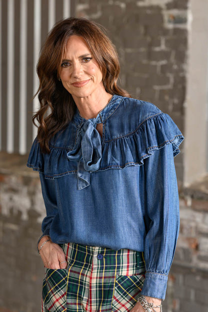 A woman wearing a blue denim long-sleeve blouse with ruffled shoulders and a tie neckline, paired with green plaid pants, standing against a rustic brick wall background.