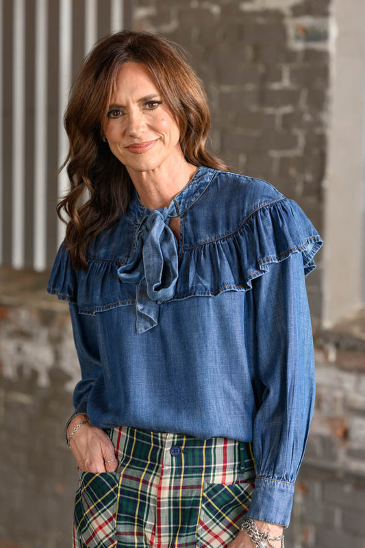 A woman wearing a blue denim long-sleeve blouse with ruffled shoulders and a tie neckline, paired with green plaid pants, standing against a rustic brick wall background.