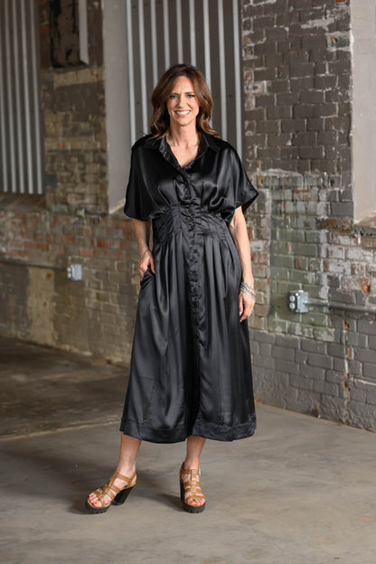 A woman wearing a black satin midi dress with short sleeves and a gathered waist, standing in an industrial brick alleyway.